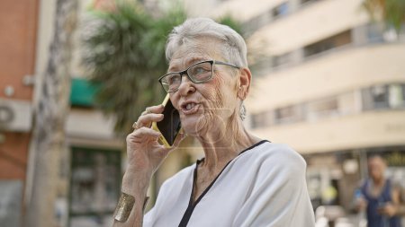 Téléchargez les photos : Senior femme aux cheveux gris maîtrise la technologie moderne, absorbé dans une conversation téléphonique sérieuse sur la rue ensoleillée de la ville - en image libre de droit
