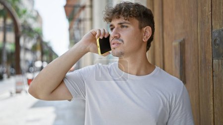 Photo for Cool-looking, handsome young hispanic man seriously engaged in conversation on his smartphone, standing under the sunny urban street's casual fashion backdrop with a concentrated expression. - Royalty Free Image