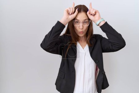 Téléchargez les photos : Belle femme brune portant une veste d'affaires et des lunettes faisant un geste drôle avec le doigt sur la tête comme des cornes de taureau - en image libre de droit