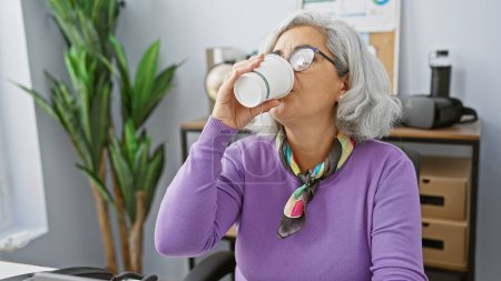 Photo for A mature woman sips coffee in a modern office setting, reflecting a casual professional break moment. - Royalty Free Image