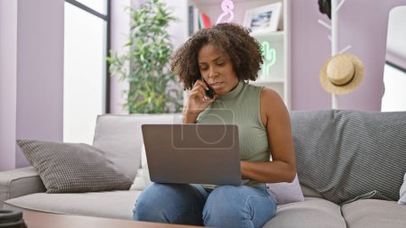 Téléchargez les photos : Femme afro-américaine avec des tresses utilisant un ordinateur portable et un téléphone sur un canapé à l'intérieur. - en image libre de droit