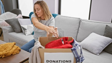 Photo for Attractive, young blonde woman sitting comfortably on her living room sofa, seriously packing clothes for charity into a package at home - Royalty Free Image