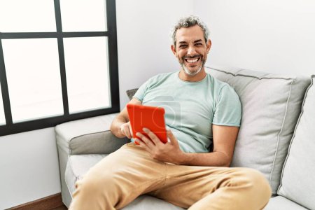 Téléchargez les photos : Moyen-âge homme aux cheveux gris utilisant pavé tactile assis sur le canapé à la maison - en image libre de droit