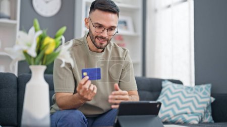 Foto de Joven hombre hispano de compras con touchpad y tarjeta de crédito sentado en el sofá en casa - Imagen libre de derechos