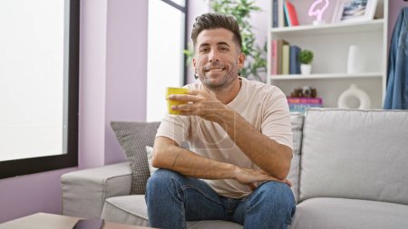 Photo for Confident young hispanic man enjoying his morning espresso, sitting comfortably on his living room sofa, a radiant smile blooming over his mug - Royalty Free Image