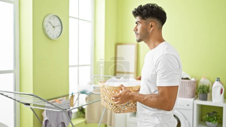 Photo for Handsome hispanic man holding laundry basket in a bright green room with washer and drying rack - Royalty Free Image