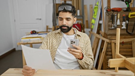Photo for Handsome hispanic man with beard using smartphone in a woodworking workshop - Royalty Free Image