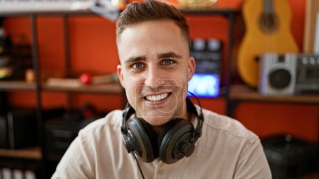 Photo for Smiling young man with headphones in music studio surrounded by musical instruments and audio equipment. - Royalty Free Image