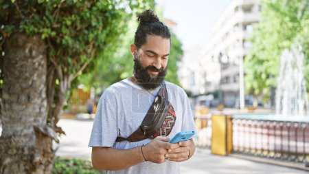 Photo for Handsome bearded hispanic man smiling while using smartphone in sunny city park - Royalty Free Image