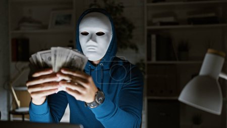 Maskierter Mann in Kapuzenpullover, der uns nachts in einem schwach beleuchteten Büro Dollar hält, was auf illegale Finanzaktivitäten hindeutet.