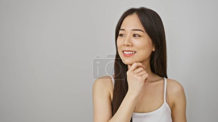 Foto de Una mujer asiática reflexiva en una camiseta blanca posa sobre un fondo blanco aislado, encarnando elegancia casual. - Imagen libre de derechos