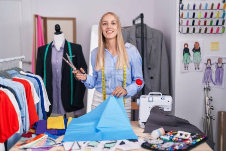 Photo for Young blonde woman tailor smiling confident cutting cloth at tailor shop - Royalty Free Image