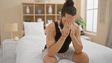 Photo for A distressed hispanic man sitting on a bedroom bed, covering his face with hands in an indoor setting. - Royalty Free Image