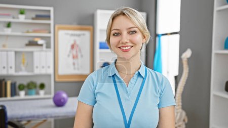 Foto de Una joven profesional con una sonrisa amistosa usando un polo azul se levanta con confianza en una clínica de fisioterapia bien equipada. - Imagen libre de derechos