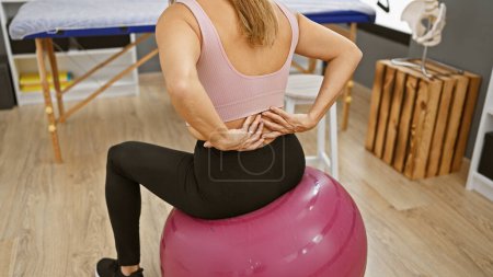 Téléchargez les photos : Une jeune femme hispanique ressent des maux de dos alors qu'elle est assise sur une balle de fitness à l'intérieur d'une clinique de désintox. - en image libre de droit