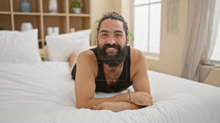 Foto de Un alegre hombre hispano barbudo relajándose en un dormitorio moderno, exudando un ambiente informal y acogedor. - Imagen libre de derechos