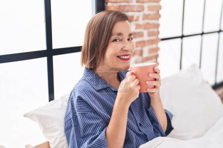 Téléchargez les photos : Femme d'âge moyen buvant une tasse de café assis sur le lit dans la chambre - en image libre de droit