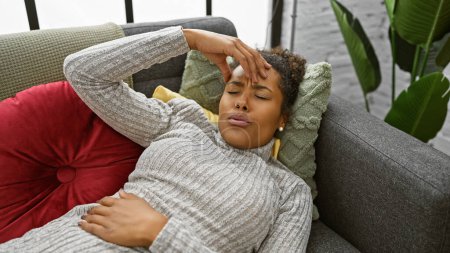 Young woman feeling unwell, lies on couch at home with hand on forehead.