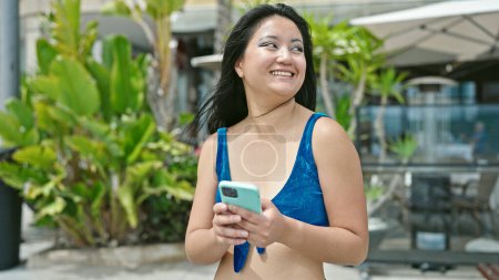 Téléchargez les photos : Jeune femme chinoise touriste portant un bikini en utilisant smartphone à la rue - en image libre de droit