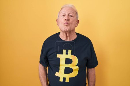 Téléchargez les photos : Homme âgé avec les cheveux gris portant Bitcoin t-shirt regardant la caméra souffler un baiser sur l'air étant belle et sexy. expression d'amour. - en image libre de droit