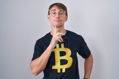 Téléchargez les photos : Blanc blond homme portant Bitcoin t-shirt pensée concentré sur le doute avec le doigt sur le menton et la recherche jusqu'à se demander - en image libre de droit