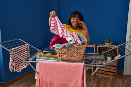 Photo for Young beautiful latin woman talking on smartphone hanging clothes on clothesline at laundry room - Royalty Free Image