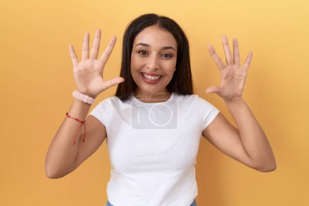 Téléchargez les photos : Jeune femme arabe portant casual t-shirt blanc sur fond jaune montrant et pointant vers le haut avec les doigts numéro dix tout en souriant confiant et heureux. - en image libre de droit