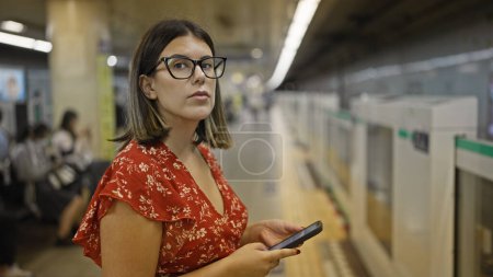 Superbe femme hispanique, lunettes allumées, debout à la gare souterraine, anticipant son voyage dans le métro, absorbée par son téléphone
