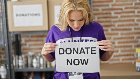 Foto de Joven mujer rubia voluntaria sosteniendo donar ahora pancarta en el centro de caridad - Imagen libre de derechos