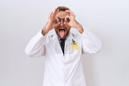 Foto de Médico de mediana edad con barba usando un abrigo blanco haciendo un buen gesto como binoculares sacando la lengua, ojos mirando a través de los dedos. expresión loca. - Imagen libre de derechos