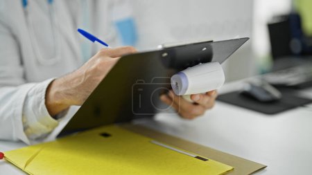 Foto de Joven médico hispano escribiendo informe médico sosteniendo termómetro en la clínica - Imagen libre de derechos