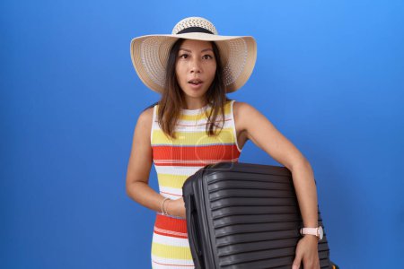 Téléchargez les photos : Femme chinoise du Moyen Âge tenant une valise allant en vacances d'été effrayée et étonnée avec la bouche ouverte pour la surprise, visage incrédule - en image libre de droit