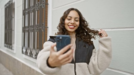 Photo for A smiling young woman takes a selfie on a city street, showcasing modern urban lifestyle and technology use. - Royalty Free Image