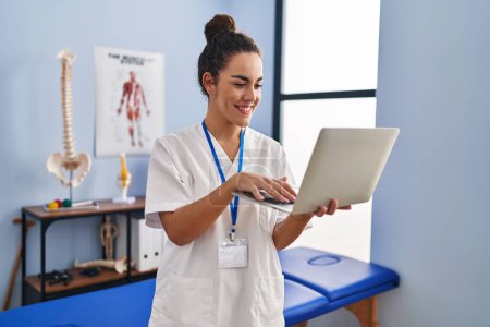 Foto de Young beautiful hispanic woman physiotherapist smiling confident using laptop at rehab clinic - Imagen libre de derechos