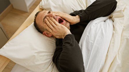 Téléchargez les photos : Un jeune homme en détresse couché à l'intérieur du lit couvre son visage de ses mains, montrant des signes de tristesse ou d'épuisement. - en image libre de droit