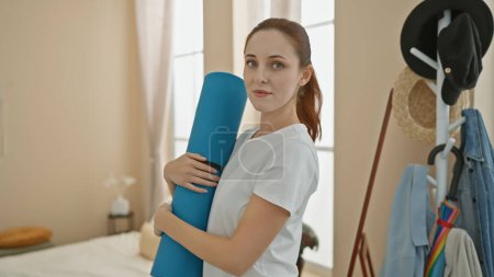 Téléchargez les photos : Une jeune femme caucasienne tient un tapis de yoga dans sa chambre sereine, décrivant un mode de vie sain. - en image libre de droit