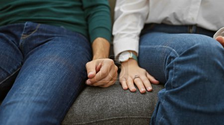 A man and woman sit closely on a couch, hands tenderly clasped, symbolizing love in an intimate indoor setting.
