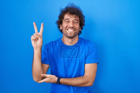 Téléchargez les photos : Jeune homme hispanique debout sur fond bleu souriant avec un sourire heureux clin d'oeil à la caméra faisant signe de victoire avec les doigts. numéro deux. - en image libre de droit