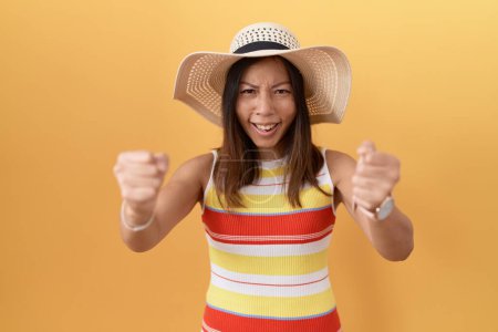 Foto de Mujer china de mediana edad con sombrero de verano sobre fondo amarillo enojado y loco levantando los puños frustrado y furioso mientras gritaba con ira. rabia y concepto agresivo. - Imagen libre de derechos