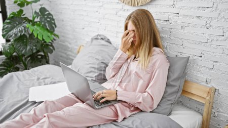 Foto de Una mujer estresada en pijama trabajando en un portátil en la cama, frotándose los ojos, con una hoja de papel a su lado en un dormitorio luminoso. - Imagen libre de derechos