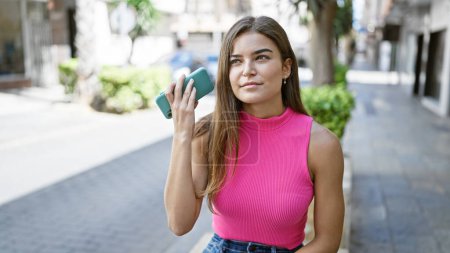 Photo for Relaxed young adult, a beautiful hispanic woman, seriously concentrates on listening to a personal message on her phone, ensconced on a city street bench. - Royalty Free Image