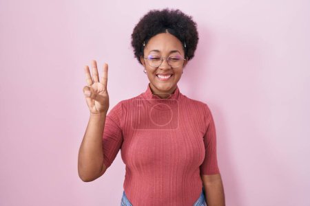 Foto de Hermosa mujer africana con el pelo rizado de pie sobre fondo rosa mostrando y apuntando hacia arriba con los dedos número tres mientras sonríe confiado y feliz. - Imagen libre de derechos
