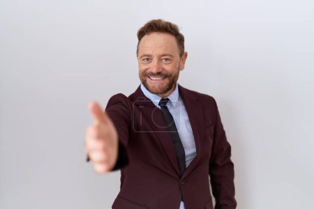 Foto de Hombre de negocios de mediana edad con barba vistiendo traje y corbata sonriente amable ofreciendo apretón de manos como saludo y bienvenida. negocios exitosos. - Imagen libre de derechos