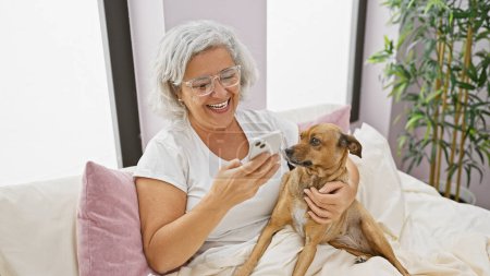 Téléchargez les photos : Une femme mûre joyeuse utilisant un smartphone câline avec son chien sur un lit à l'intérieur d'une chambre confortable. - en image libre de droit