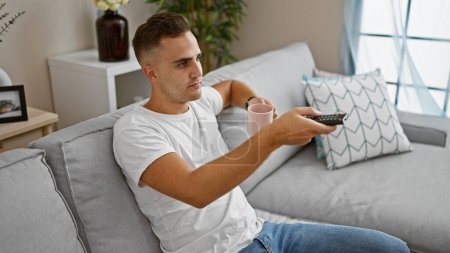 Photo for Handsome man sitting comfortably on a gray couch at home holding a pink mug and remote control - Royalty Free Image
