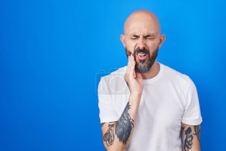 Foto de Hombre hispano con tatuajes de pie sobre fondo azul tocando la boca con la mano con expresión dolorosa debido a dolor de muelas o enfermedad dental en los dientes. dentista - Imagen libre de derechos
