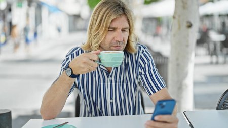 Foto de Hombre guapo y rubio bebiendo café mientras revisa el teléfono inteligente en una terraza al aire libre en la ciudad. - Imagen libre de derechos