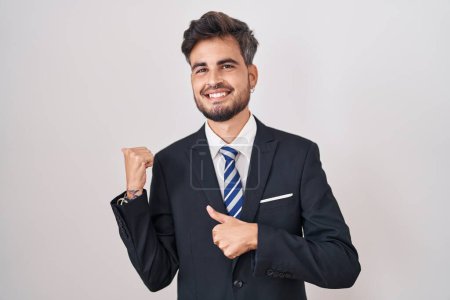 Foto de Joven hombre hispano con tatuajes con traje de negocios y corbata apuntando a la espalda con la mano y los pulgares hacia arriba, sonriendo confiado - Imagen libre de derechos