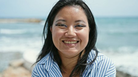 Téléchargez les photos : Jeune femme chinoise touriste souriant confiant assis sur le banc au bord de la mer - en image libre de droit