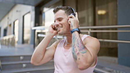 Téléchargez les photos : Homme tatoué souriant jouissant de musique avec écouteurs sur un trottoir de la ville. - en image libre de droit